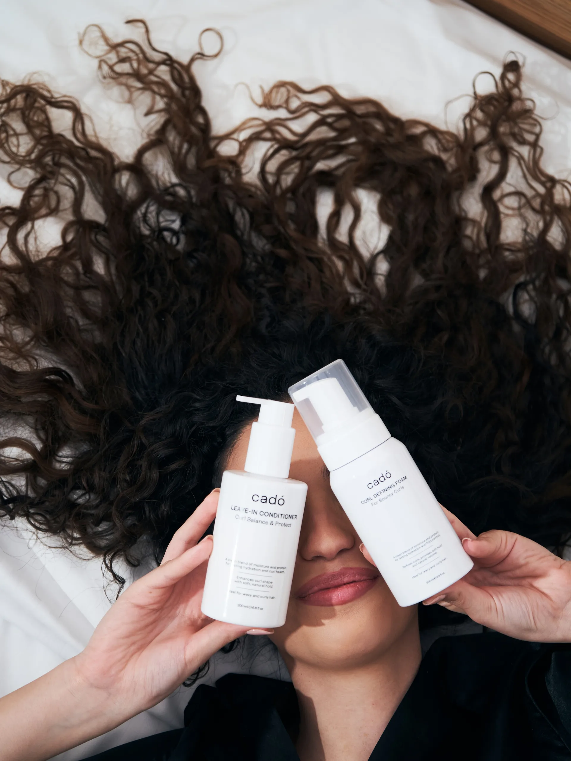 Woman with healthy defined curly hair holding cadó Leave-in Conditioner and Curl Defining Foam for professional curly hair care routine.