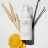 Minimalist flat lay of cadó Curl Defining Foam with natural vanilla beans, orange slices, and wheat to represent the botanical formula.