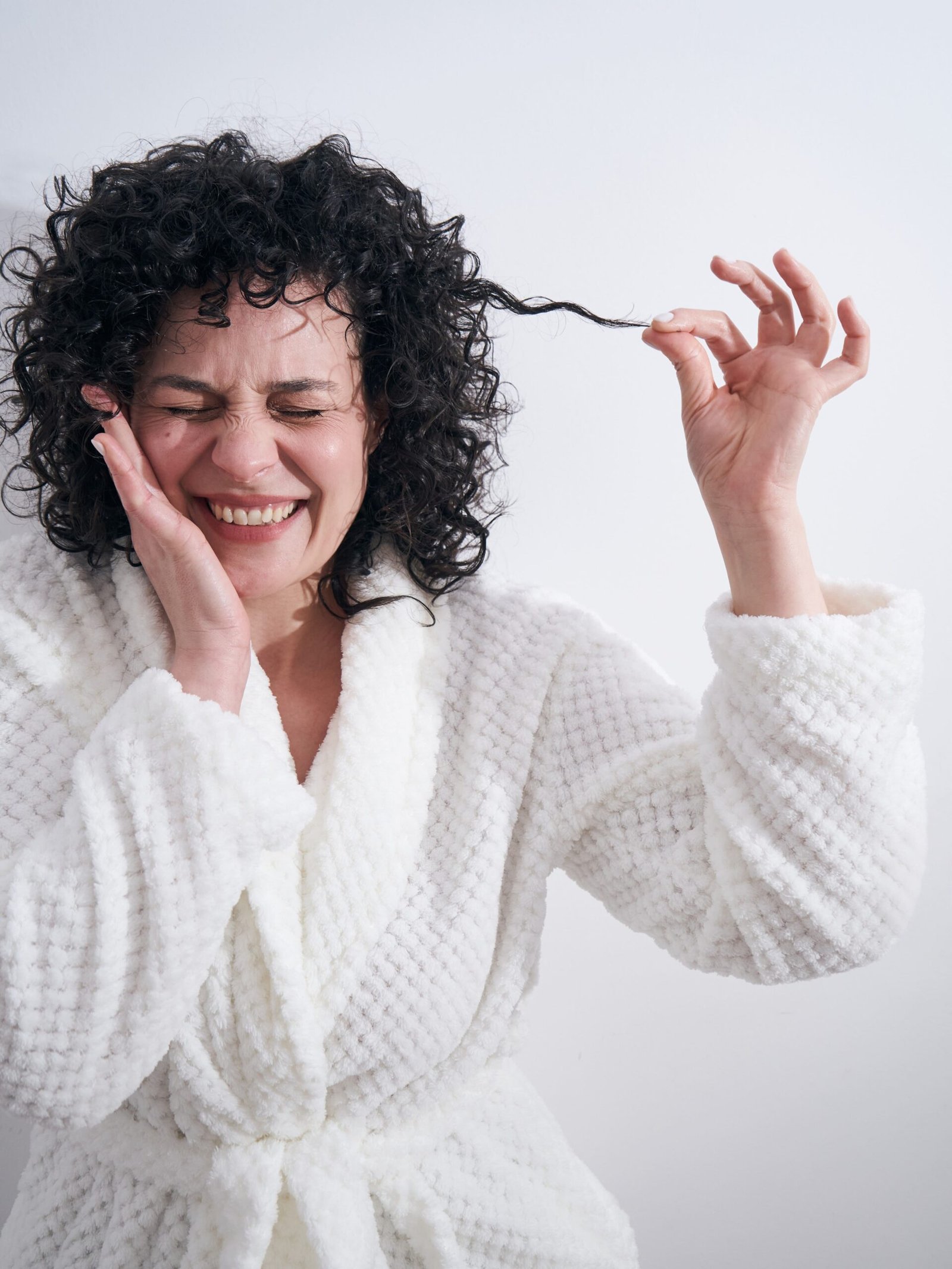 A happy woman in a white bathrobe styling her healthy, bouncy curls using cadó professional haircare.